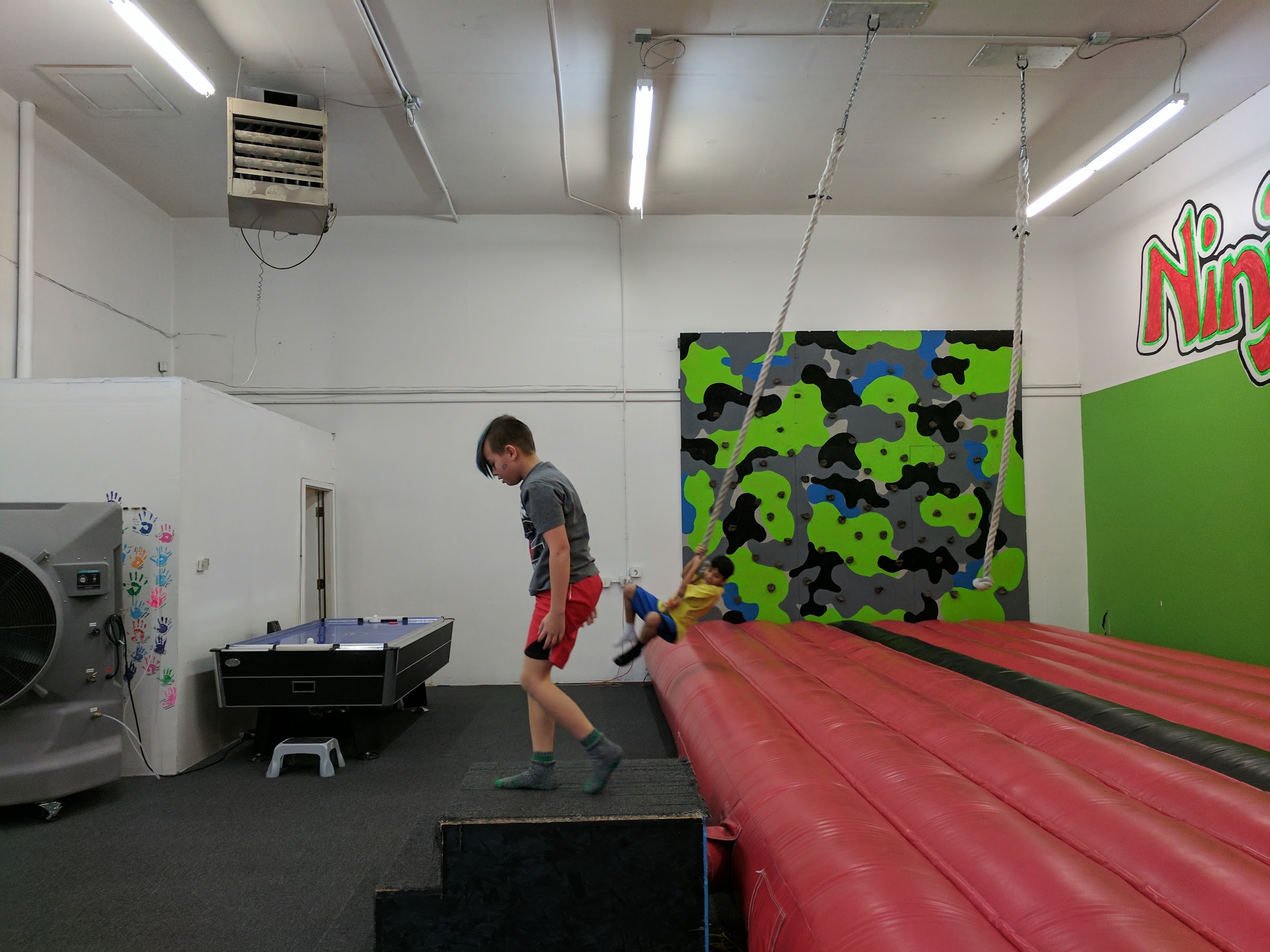 A boy on a rope swing in the ninja-style obstacle room at Inflatable Fun Gym, with a climbing wall and air-bag balance beam